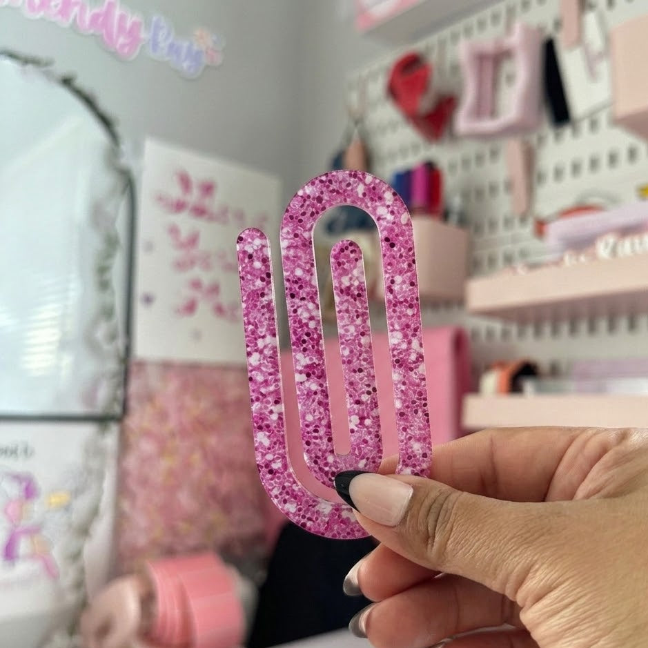 Hand holding a large pink glittery paperclip in a room with stationery items.